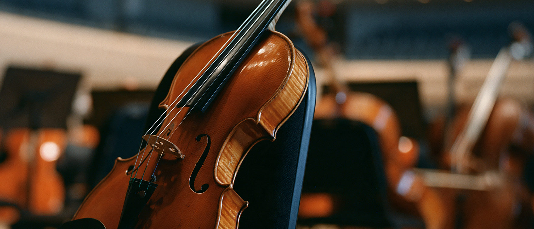 A worn golden violin resting on a black orchestra chair in an empty concert hall, instruments softly blurred behind.